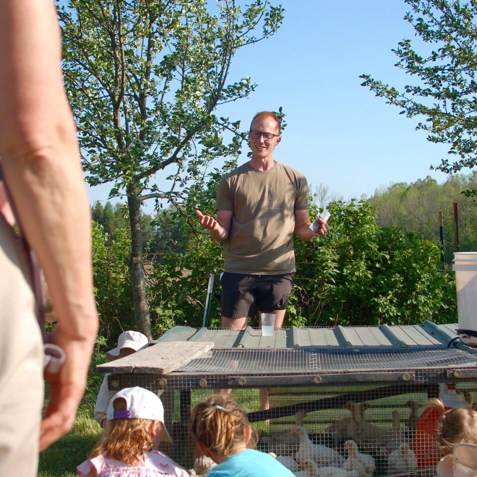 Shane standing in front of a chicken coop with children watching outdoors.