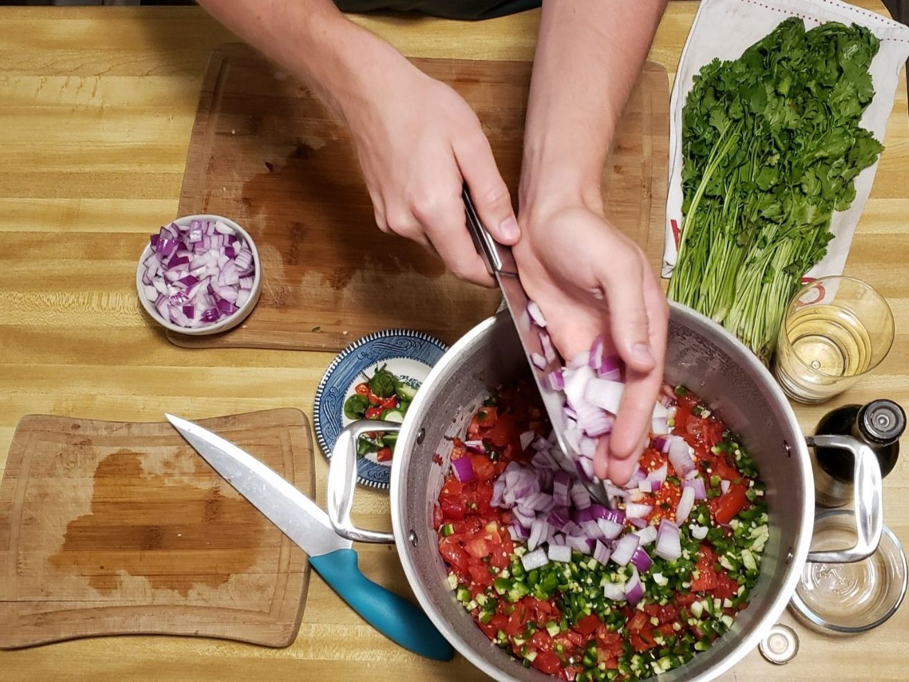 Person adding chopped onions to a pot of vegetables on a wooden table.